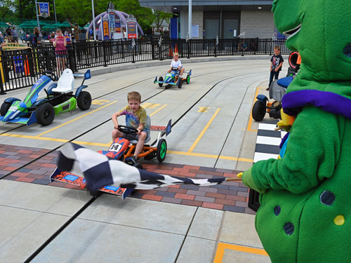 Rex the dinosaur waving a checkered racing flag while racers drive their pedal cars across a finish line that looks like the Indy 500's yard of bricks.